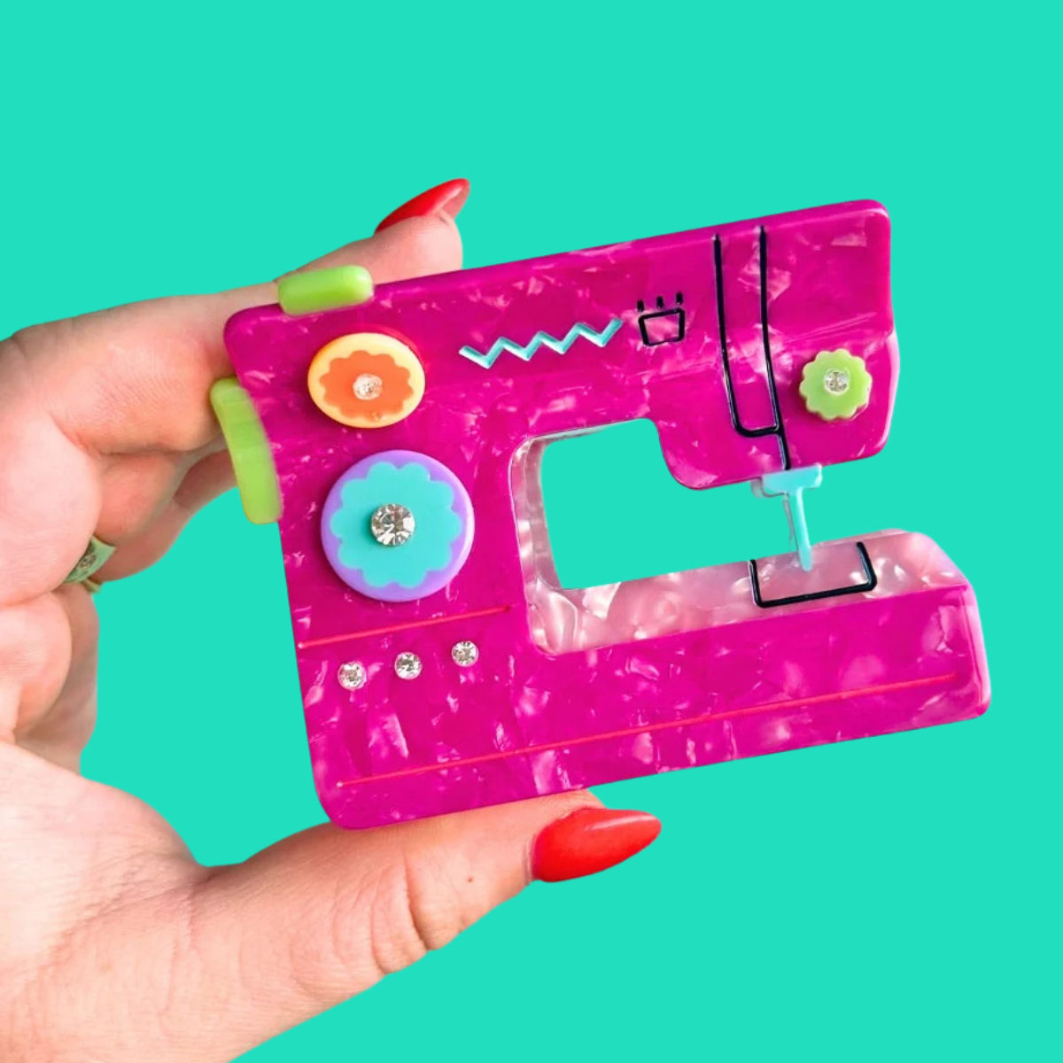 A hand with red painted nails holds a miniature pink sewing machine toy hair claw clip against a solid turquoise background. The clip features a marbled hot pink body with colorful decorative buttons, a turquoise dial, and black line-drawn details.