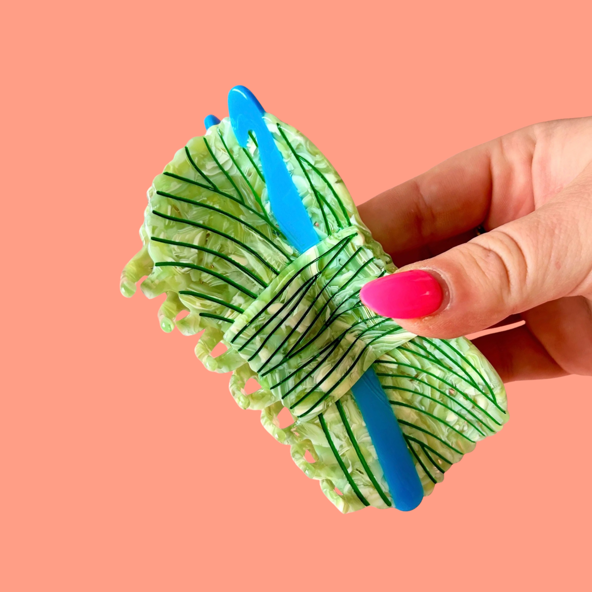 A hand with bright pink nail polish holds a light green crinkle-textured hair claw clip with dark green stripes and blue elastic bands against a solid coral pink background.