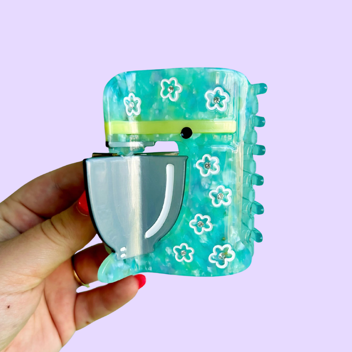 A hand holding a translucent aqua and teal stand mixer hair claw clip decorated with small white flowers and a yellow band, set against a solid lavender background.
