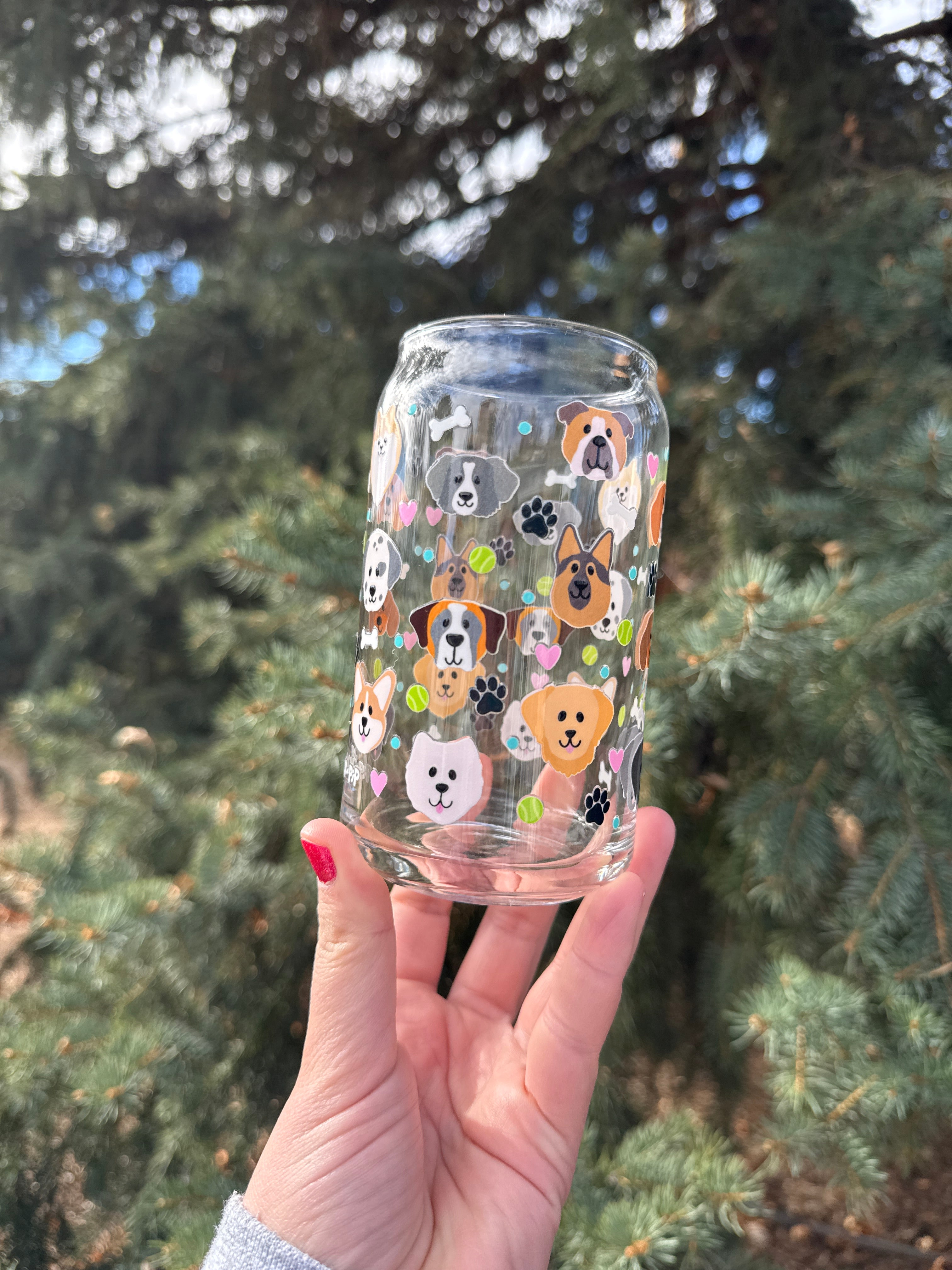 Clear glass tumbler with cartoon dog design of multiple breeds held by a hand against a tree background