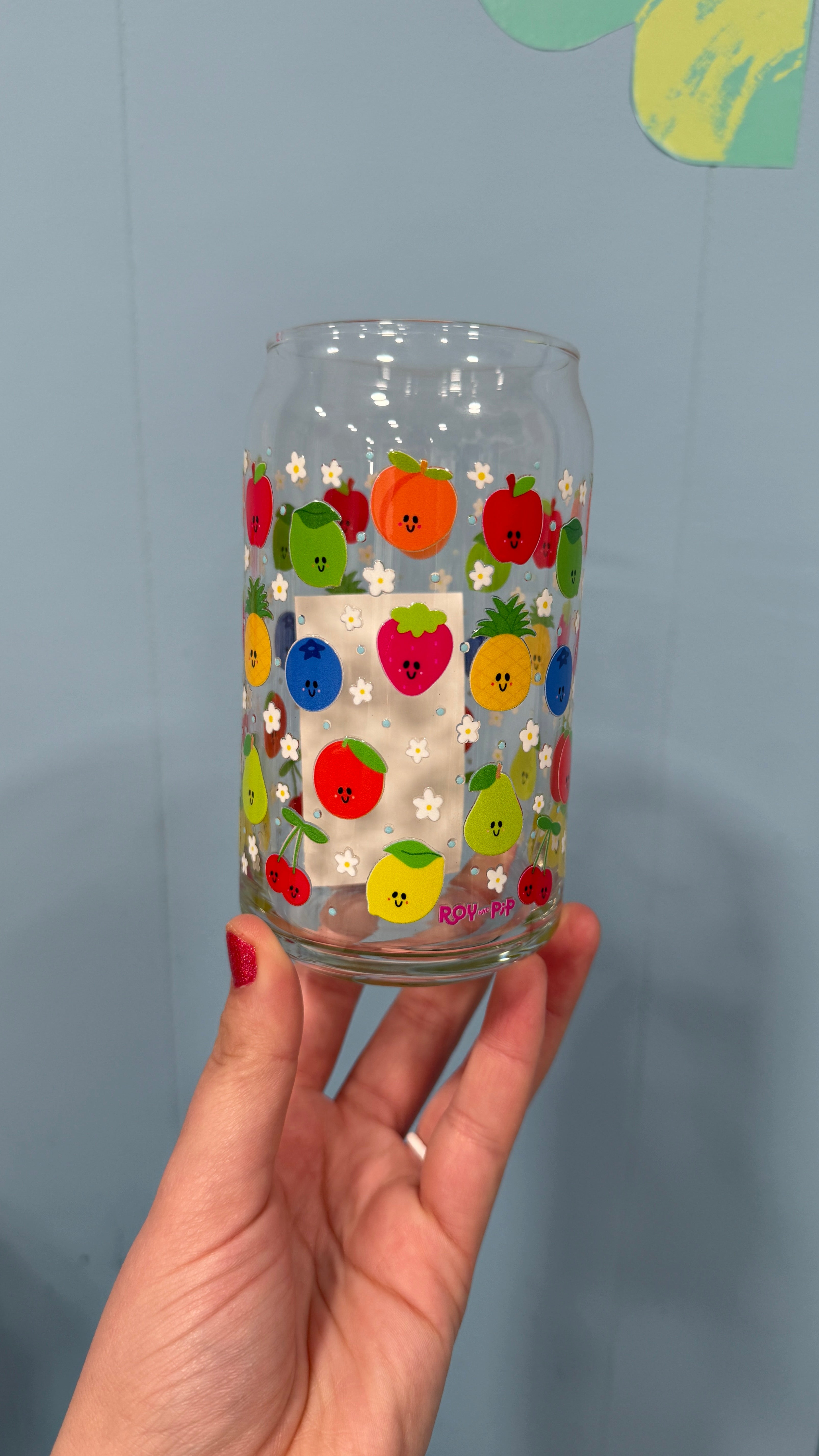 Hand holding a glass with colorful fruit design against a plain blue background