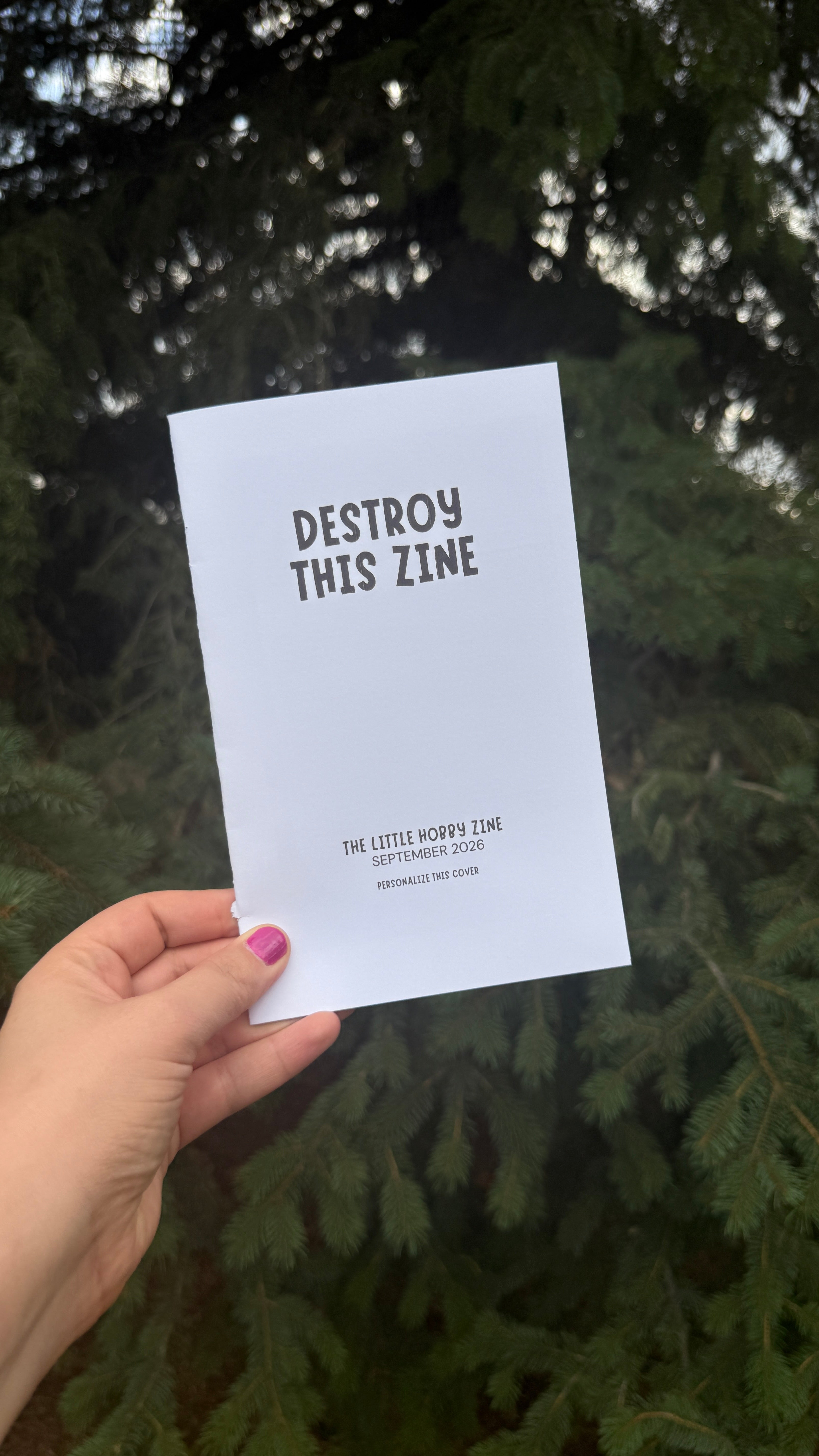 A hand with pink nail polish holds a white paper print featuring the text 'DESTROY THIS ZINE' and 'THE LITTLE WORLD ZINE SEPTEMBER 2026' against a background of dark green pine trees.