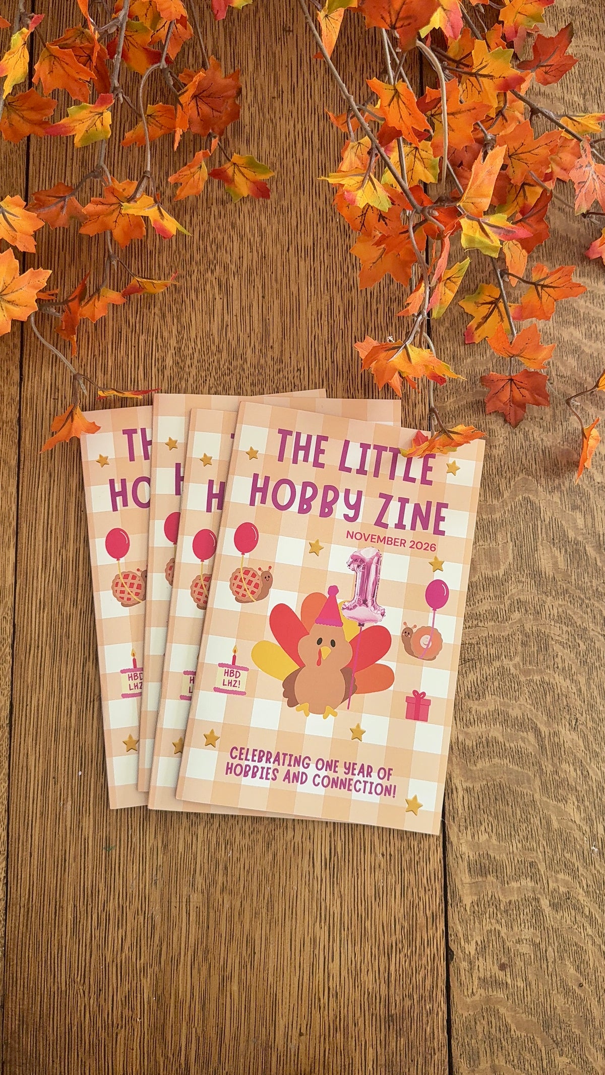 A stack of mini magazines with a turkey and snails holding balloons and ‘The Little Hobby Zine’ written across the cover, sitting on a wooden table surrounded by fall leaves.