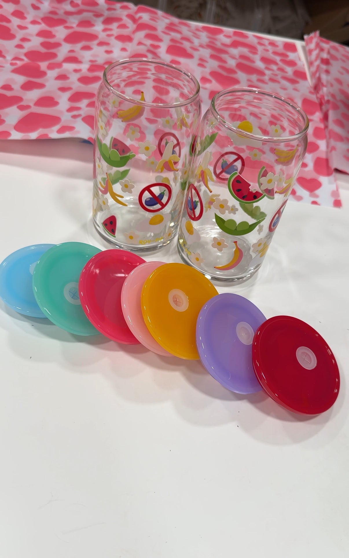 Two colorful glasses with fruit designs on a white surface, accompanied by colorful lids.