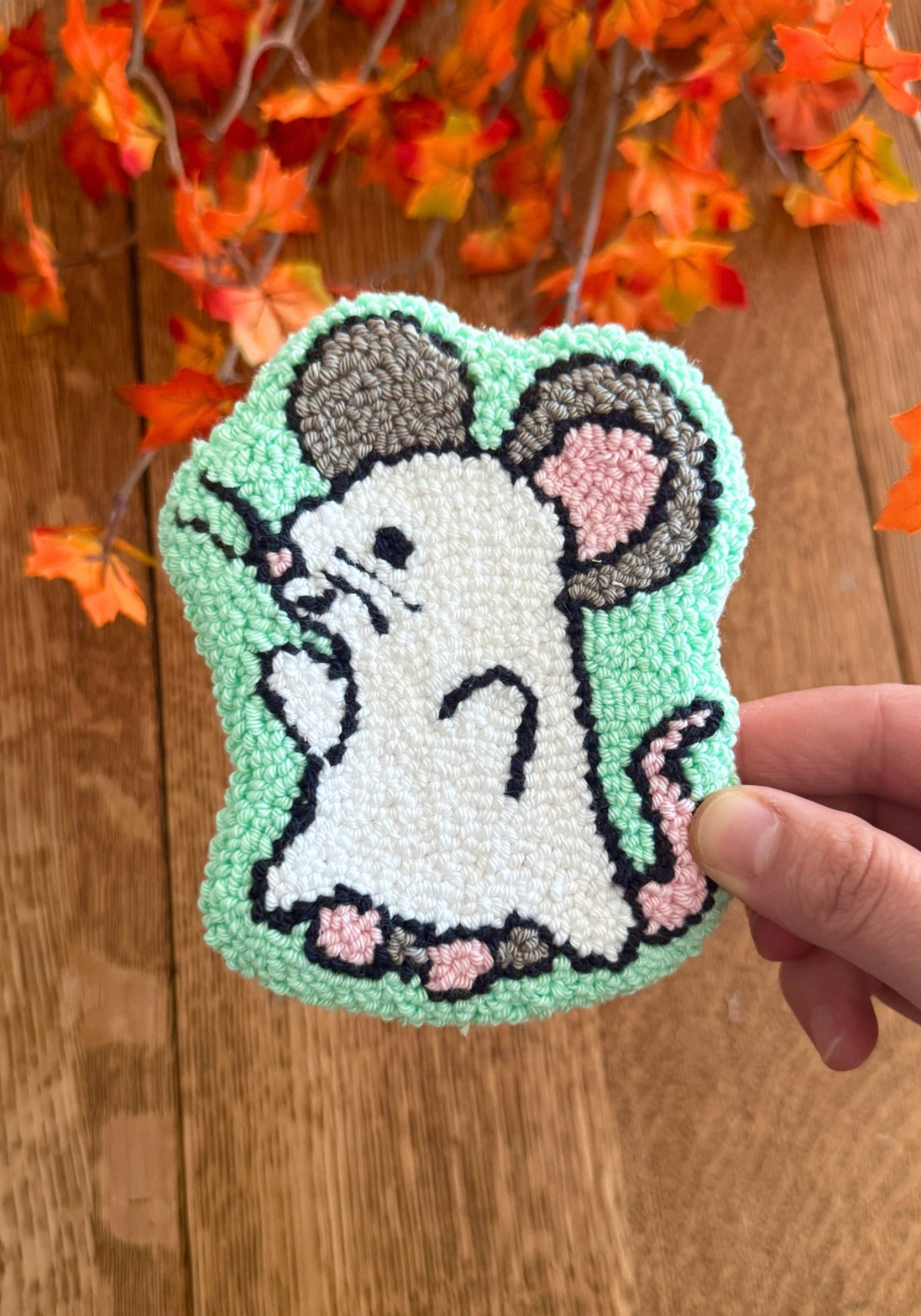 A hand holds a hand-tufted punch needle mouse ornament against a wooden background with orange autumn leaves. The mouse has a white ghost body, gray ears and tail, pink inner ears and feet, and is outlined in black on a mint green background.