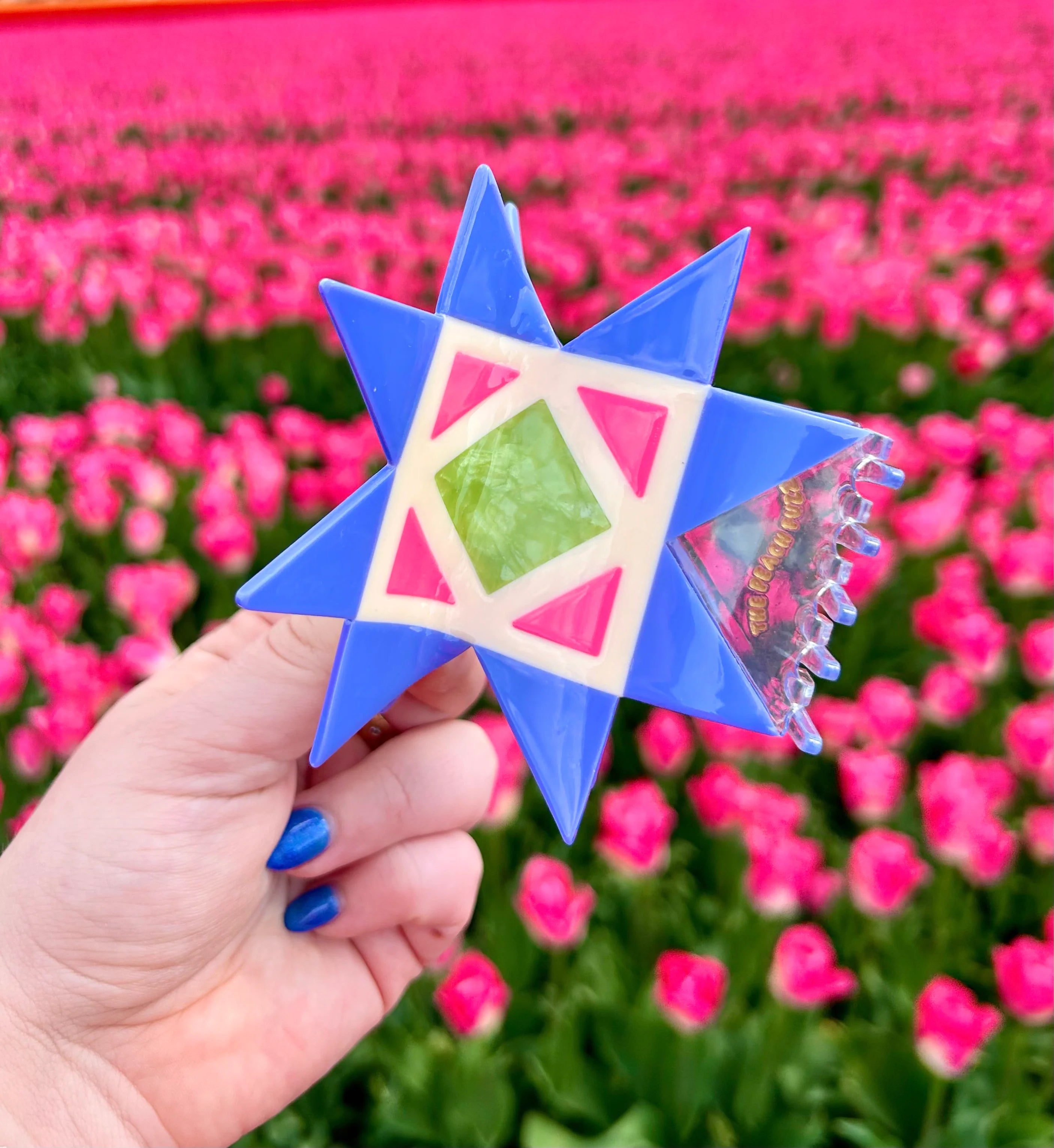 A hand with blue nail polish holds a blue and white geometric mosaic quilt block claw clip against a blurred background of a pink tulip field.