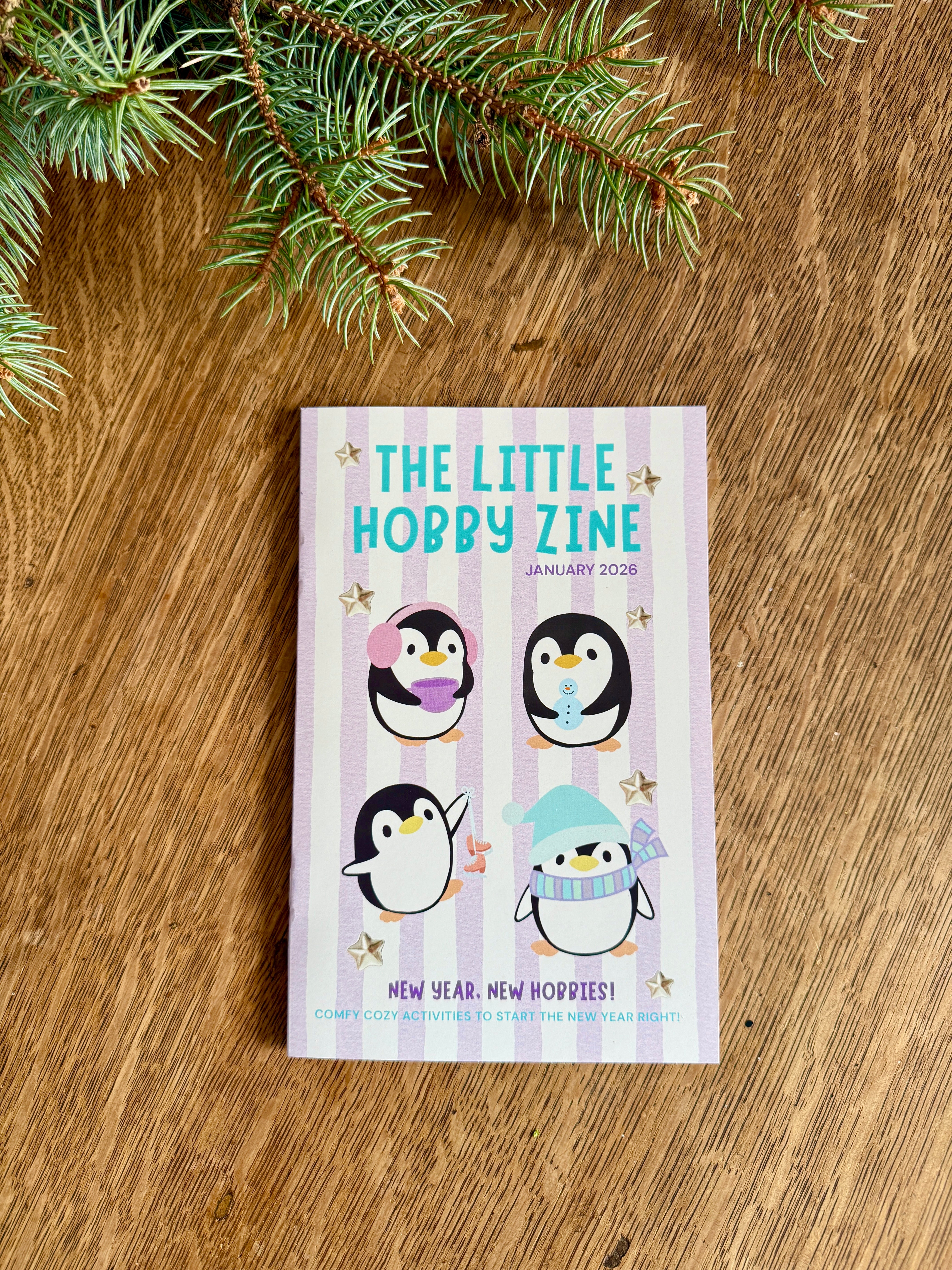 Small book titled 'The Little Hobby Zine' with penguin illustrations on a wooden surface with pine branches.