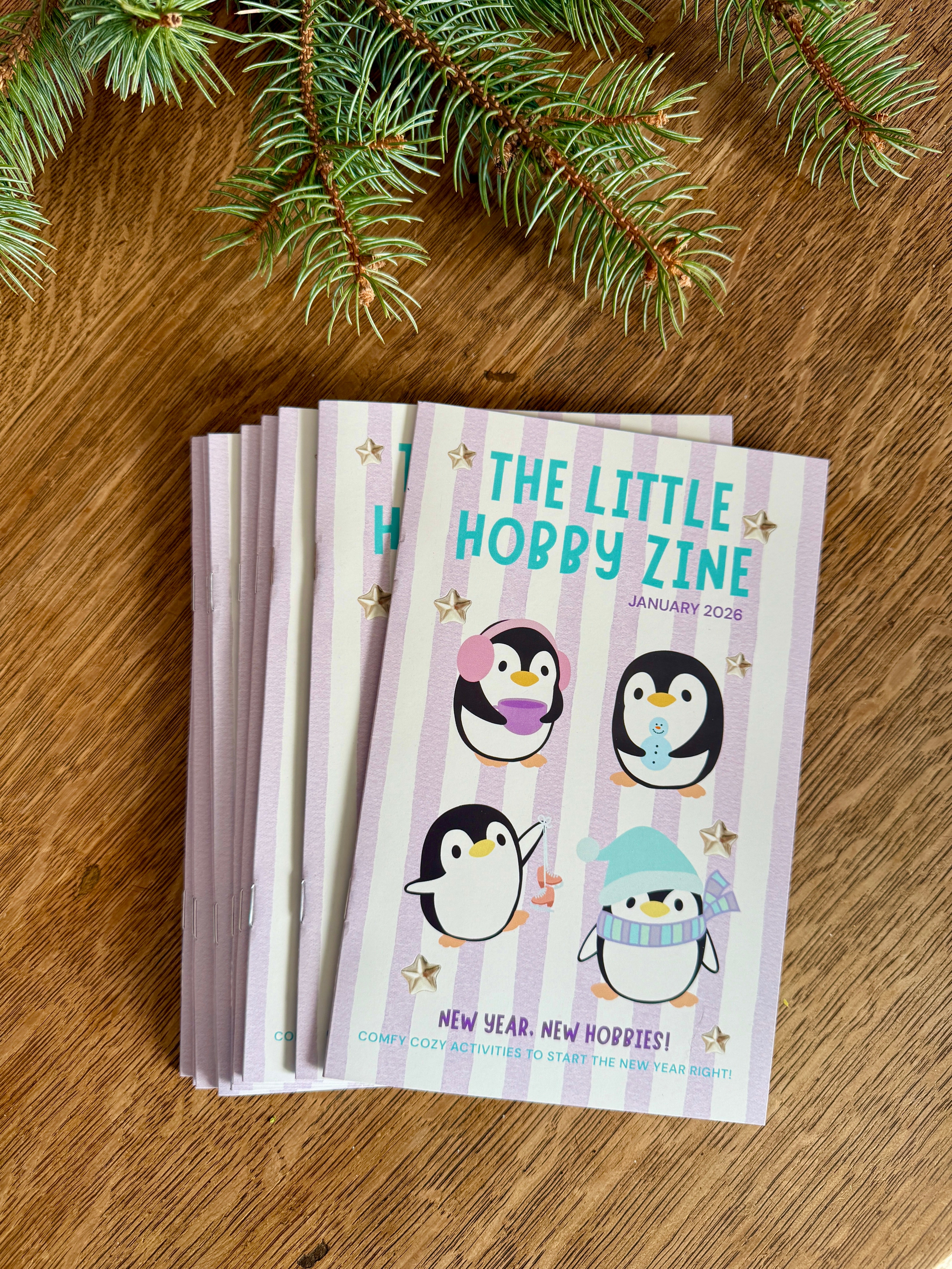 Stack of 'The Little Hobby Zine' on a wooden surface with evergreen branches.