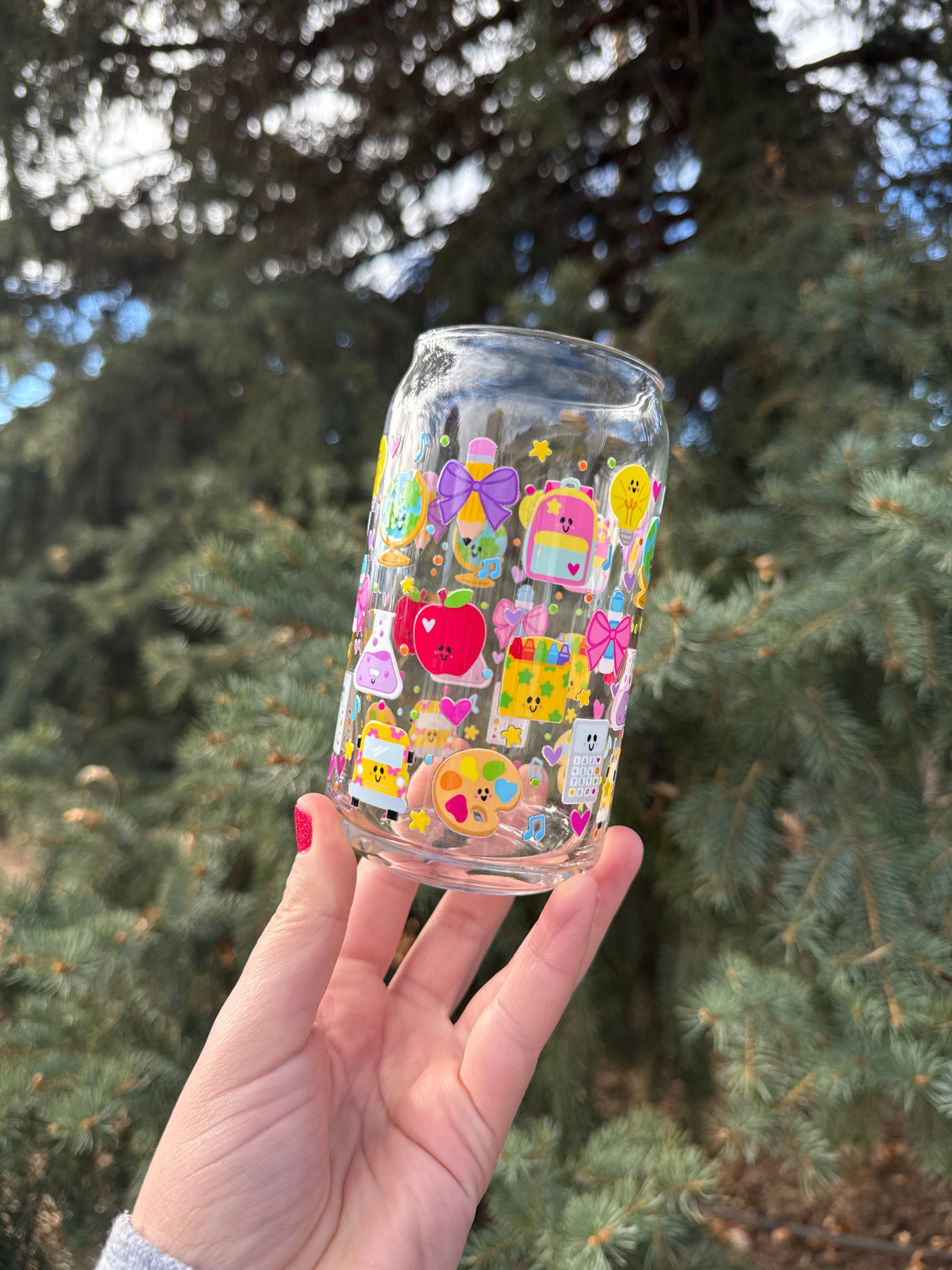 Hand holding a clear glass with colorful teacher images against a blurred green tree background