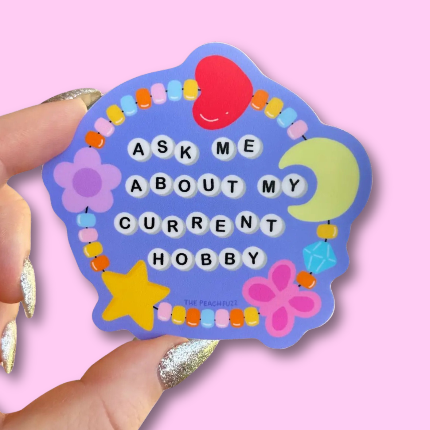 A sticker with a blue background and multicolored design, featuring text 'ASK ME ABOUT MY CURRENT HOBBY' along with a friendship bracelet illustrations such as a star, heart, and flower beads.