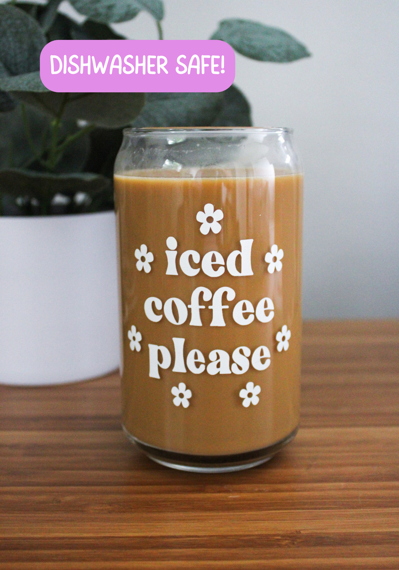 A clear glass coffee can with the text 'iced coffee please' printed on it, filled with coffee, placed on a wooden surface with a plant in the background.