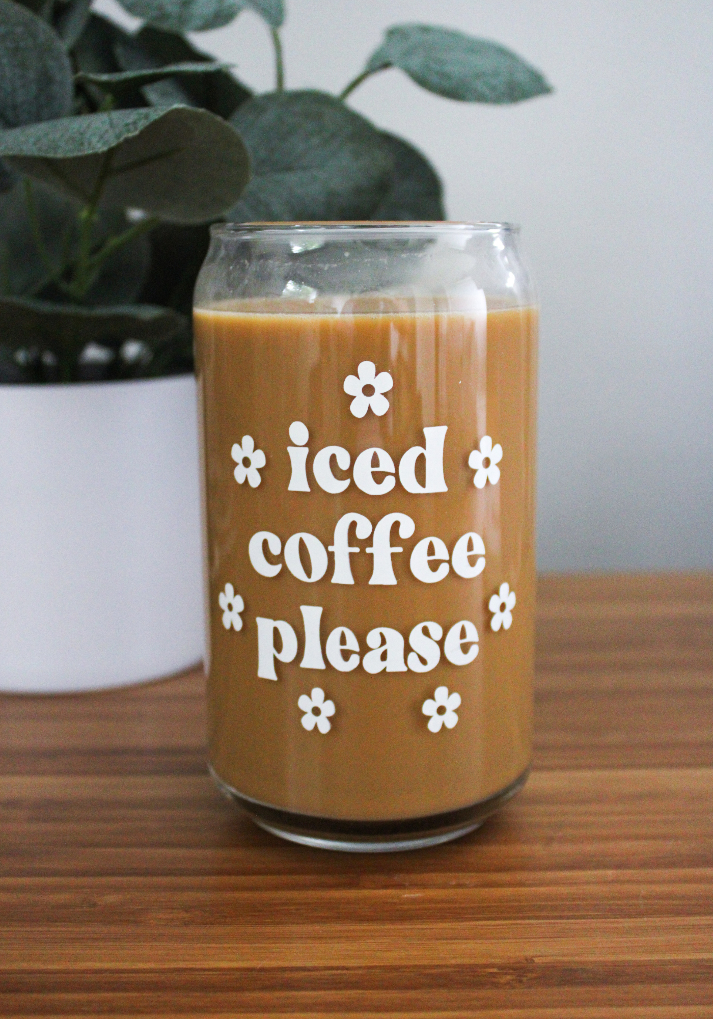 A clear glass coffee can with the text 'iced coffee please' printed on it, filled with coffee, placed on a wooden surface with a plant in the background.