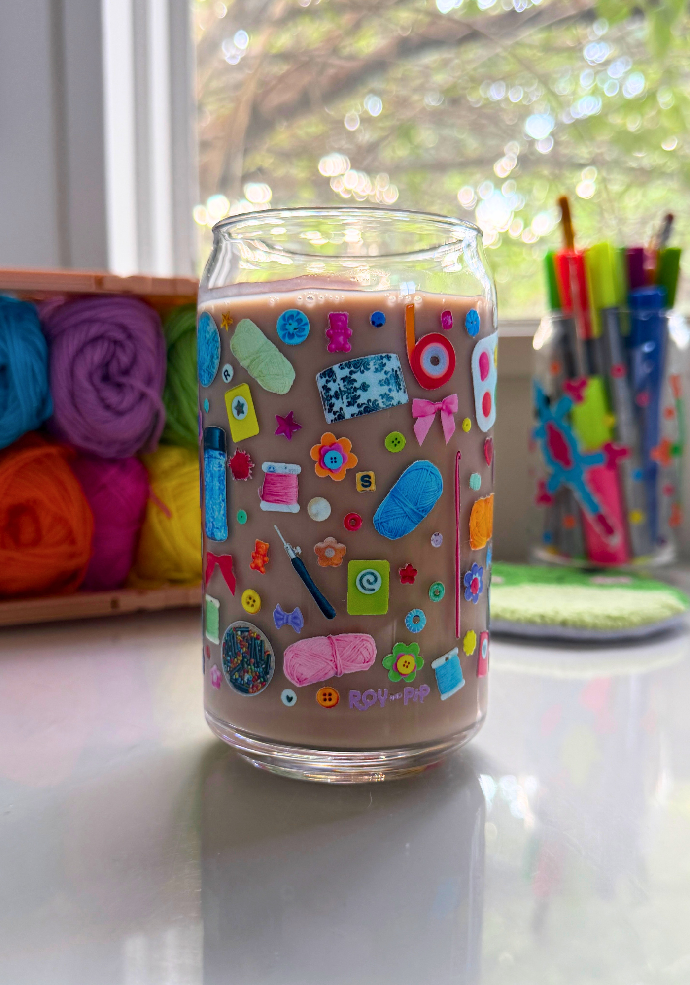 A clear glass can filled with chocolate milk, decorated with a colorful craft-themed decal, set on a reflective white surface with a window and craft supplies in the background.