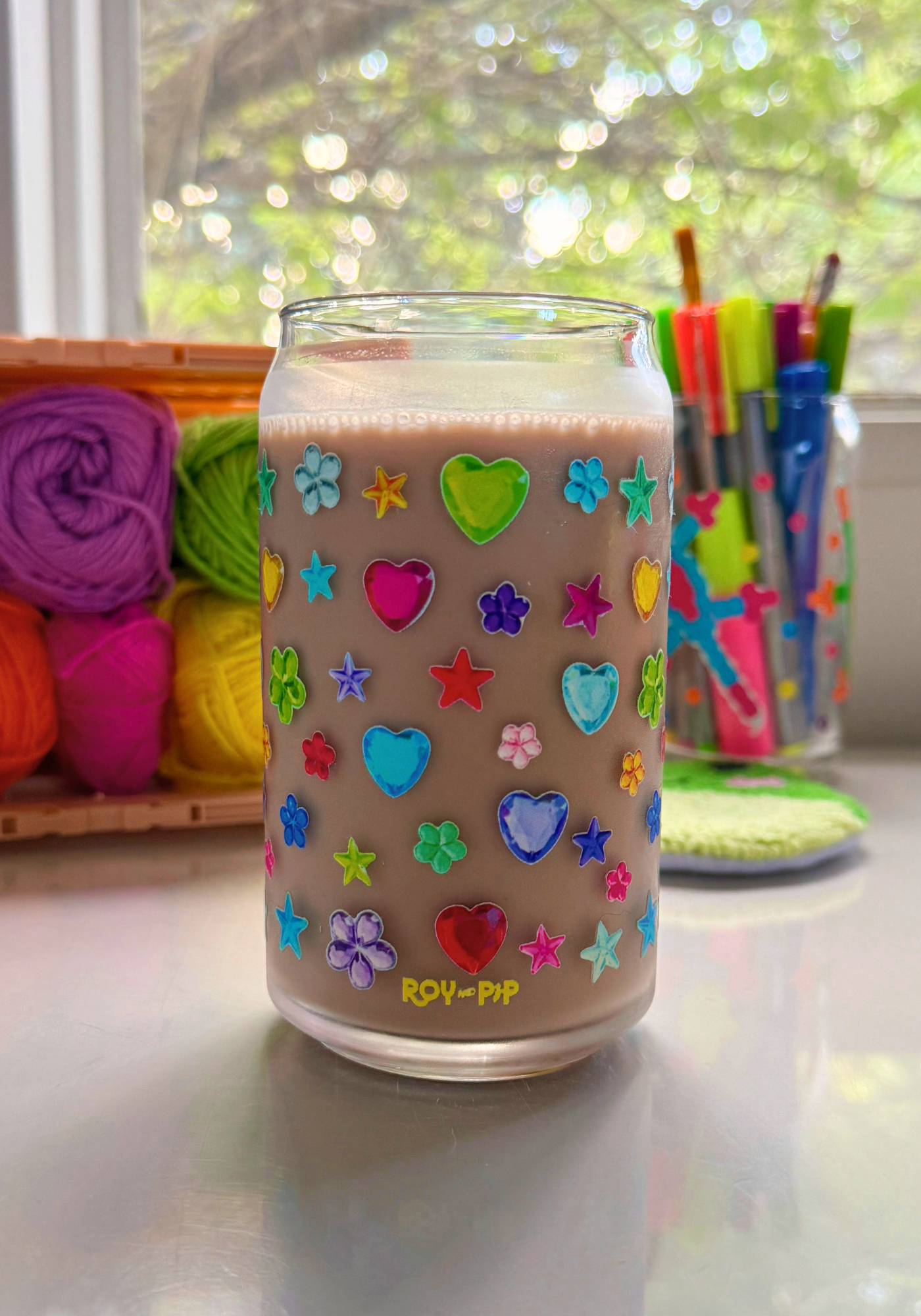 Glass can covered in a realistic colorful craft gem decal filled with chocolate milk on a desk with craft supplies.