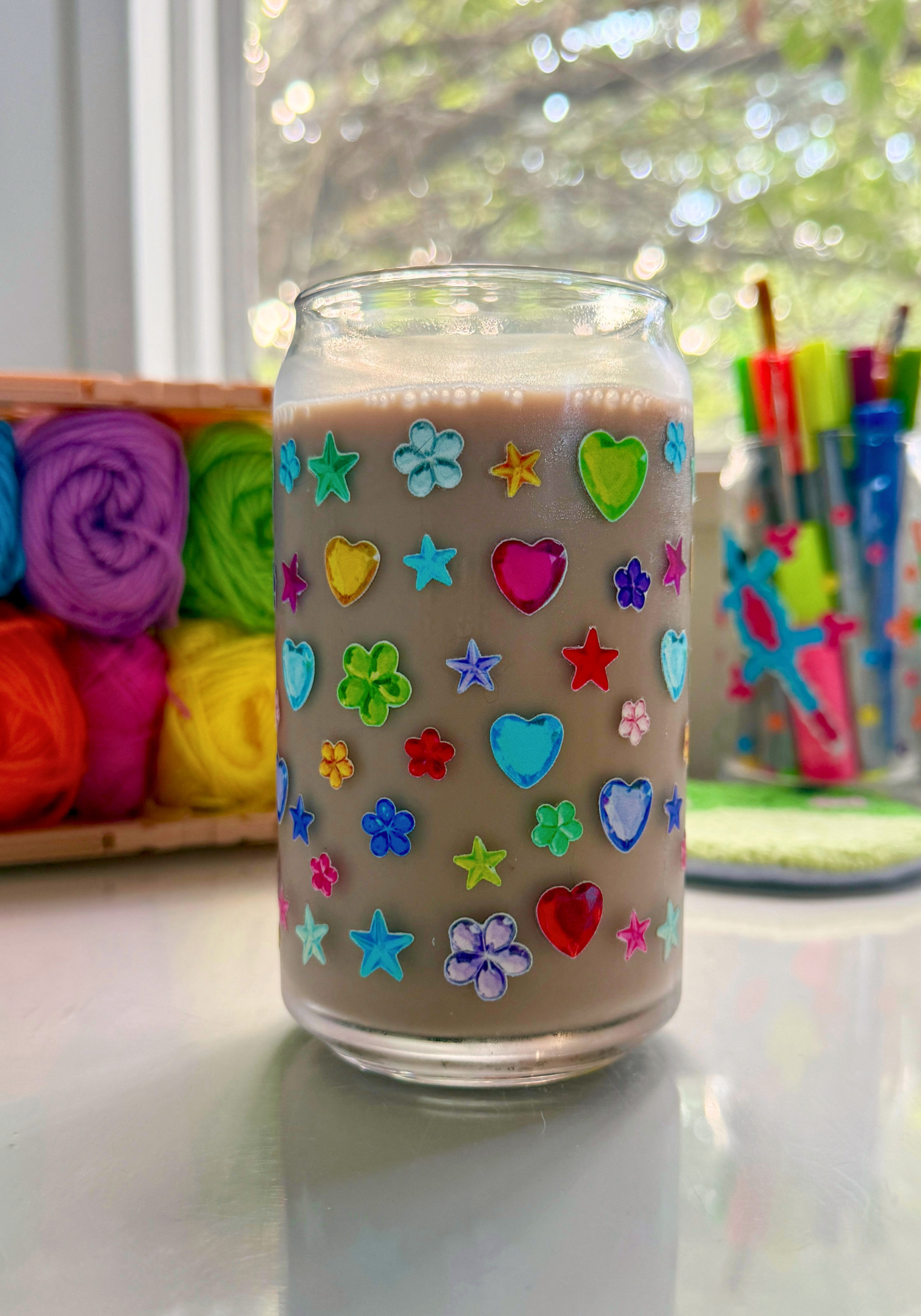 A clear glass jar filled with a light brown beverage, decorated with colorful heart and star gem sticker decals, set on a white surface with a blurred craft room background.