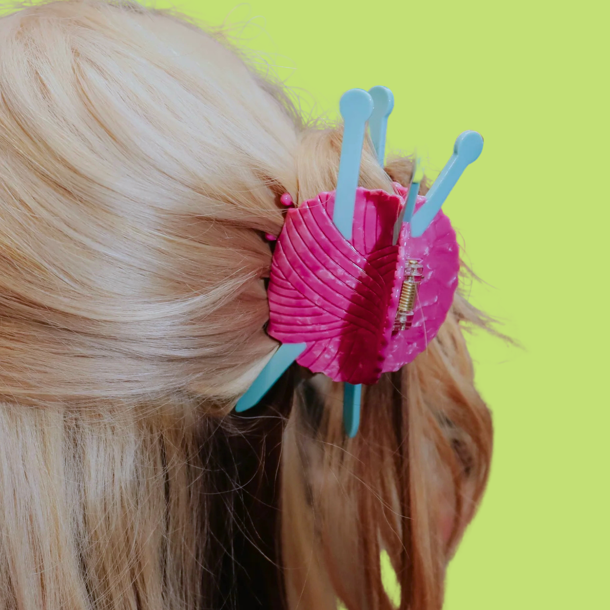 A close-up of a pink yarn and light blue knitting needle hair claw clip holding back blonde hair, shown on a bright lime green background.