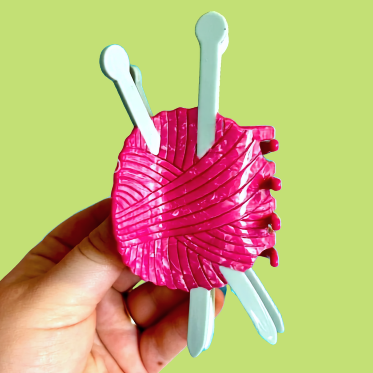 A hand holding a bright pink ball of yarn-shaped hair claw clip with four pale blue knitting needles extending from the sides, set against a solid light green background.