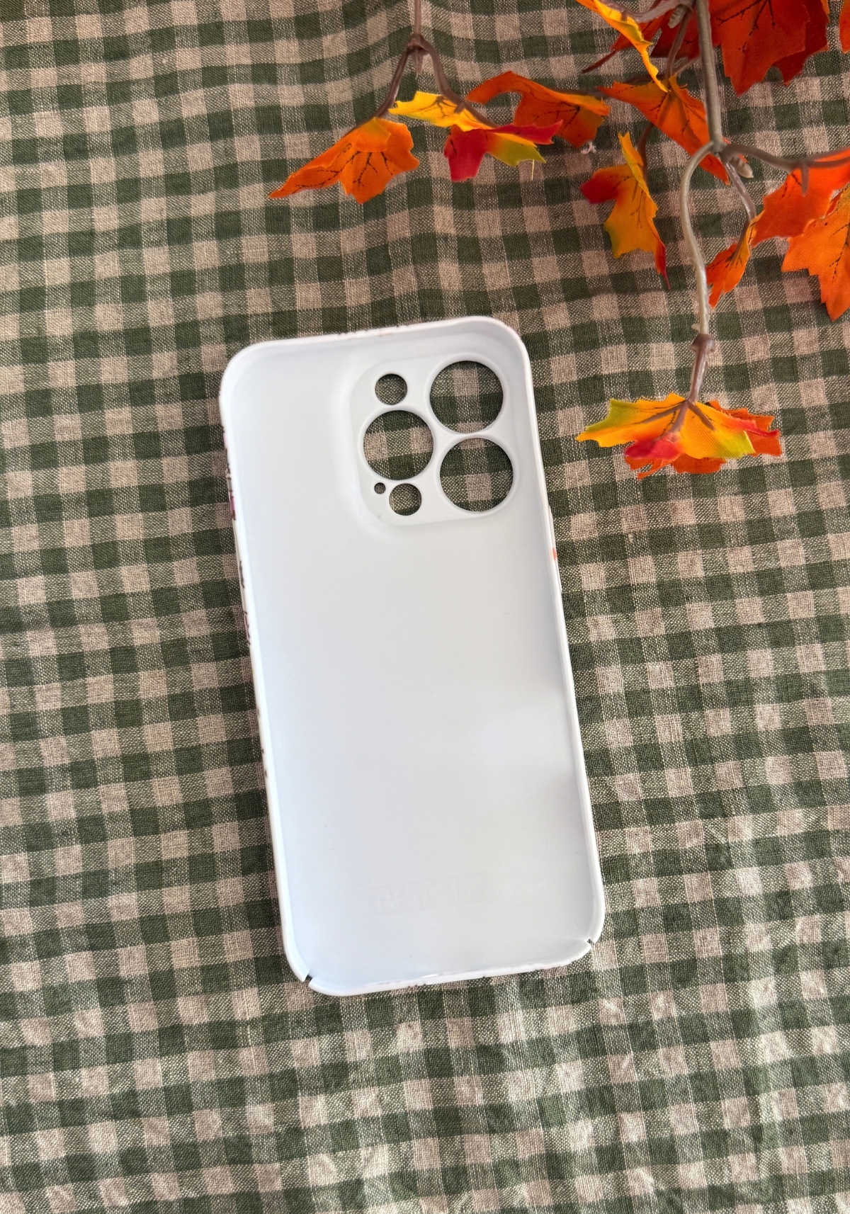 The inside of a plain acrylic white phone case on a plaid table cloth with fall leaves.