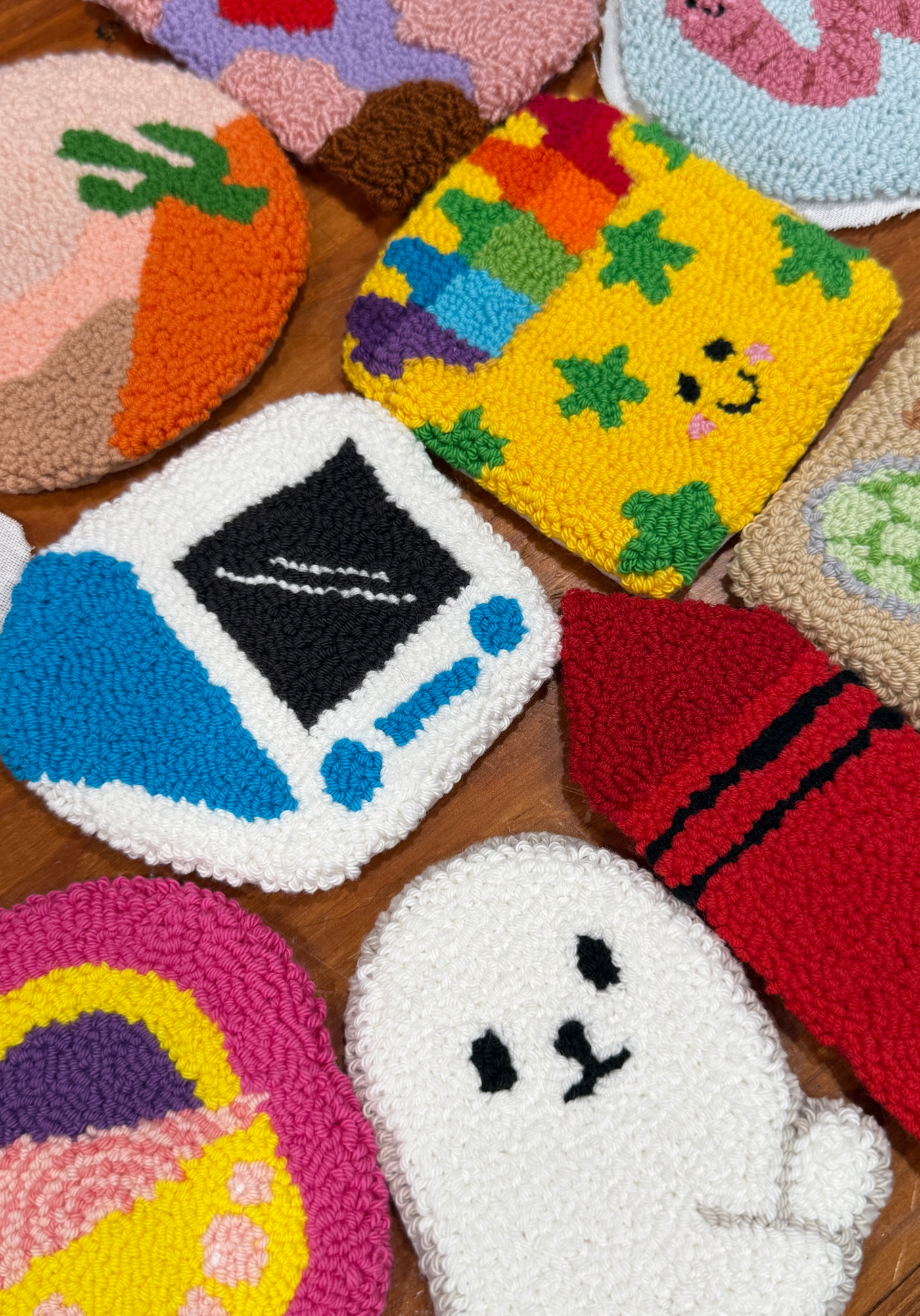 A collection of colorful, chunky tufted or looped rug coasters arranged on a wooden surface. The coasters feature whimsical cartoon designs including a retro handheld game console, a white ghost with black eyes, a red crayon, a rainbow star pattern, and a cactus.
