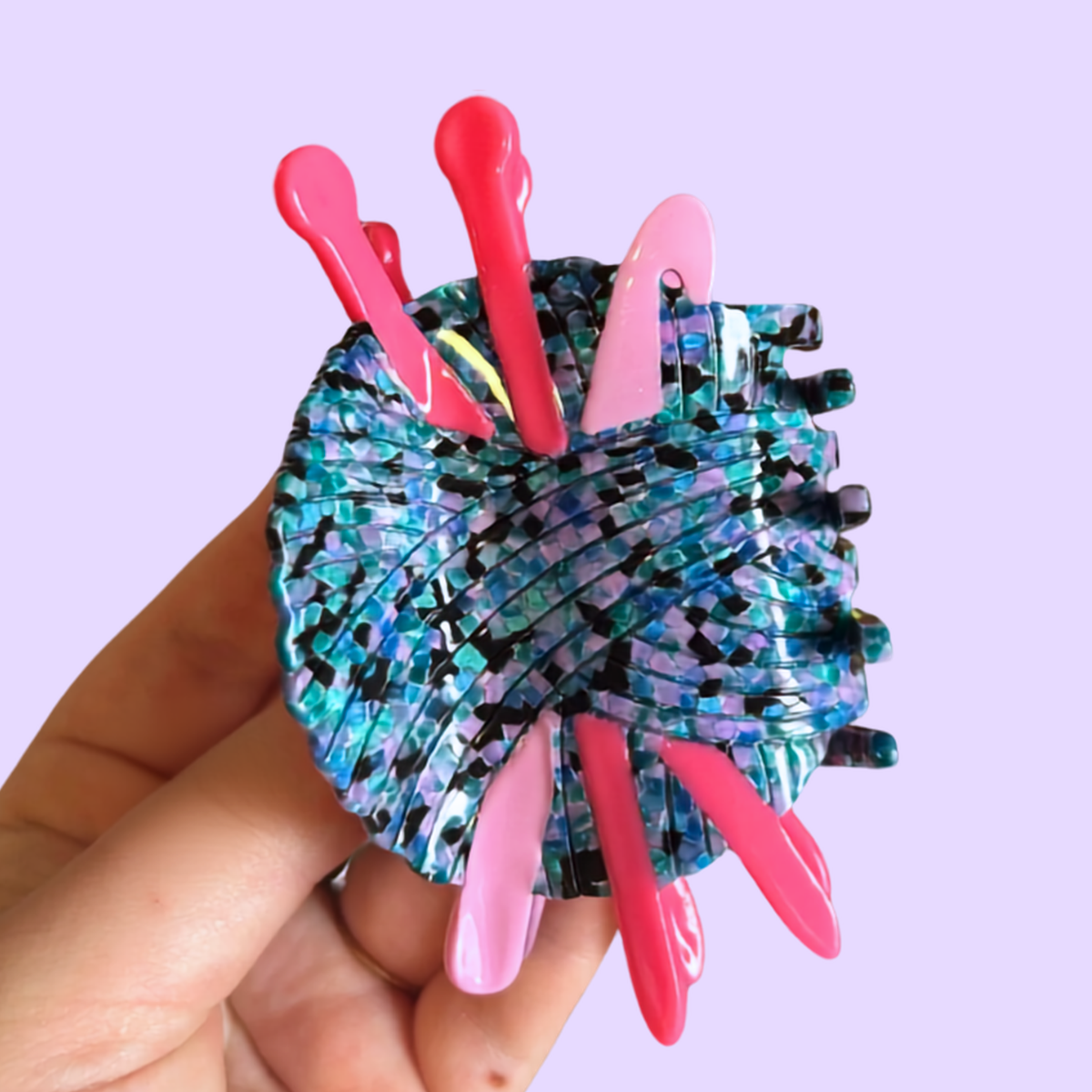 A hand holding a circular, iridescent blue and black ball of yarn hair claw with several bright pink knitting needles and crochet hook, against a solid light purple background.