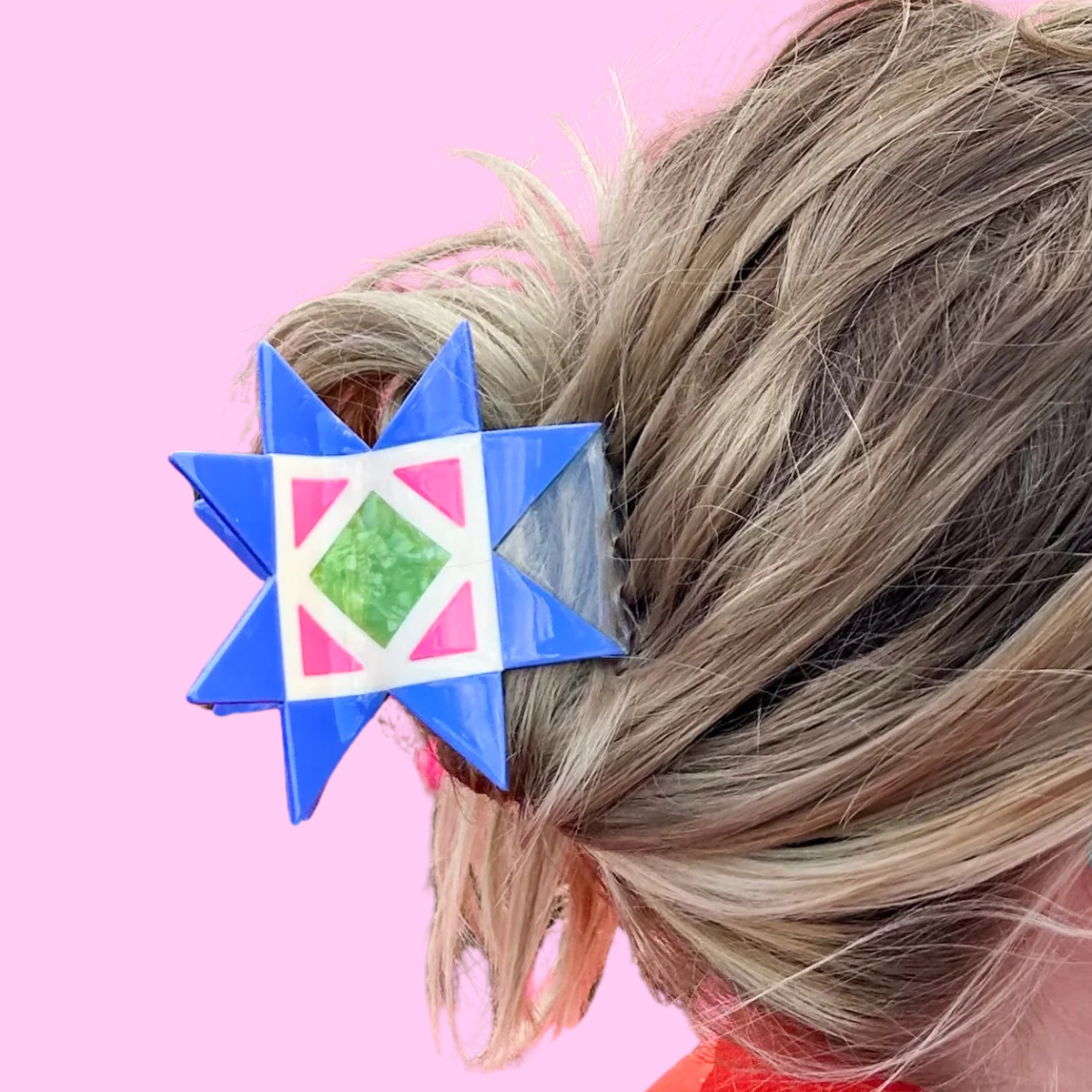 A close-up of a person's blonde hair styled in a low bun, secured with a blue mosaic quilt block hair clip featuring a pink, white, and green geometric center, set against a solid pink background.