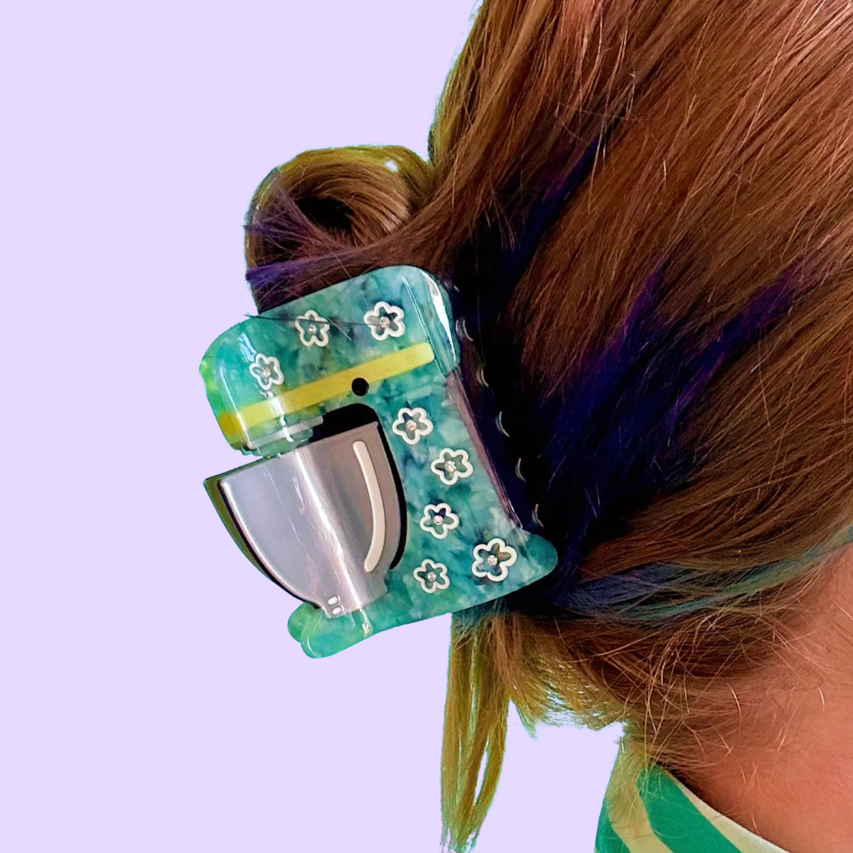 A close-up lifestyle shot of a person wearing a stand mixer hair claw clip. The clip is made of marbled teal and green acetate with white flower patterns and a yellow stripe, holding back light brown hair with purple highlights.