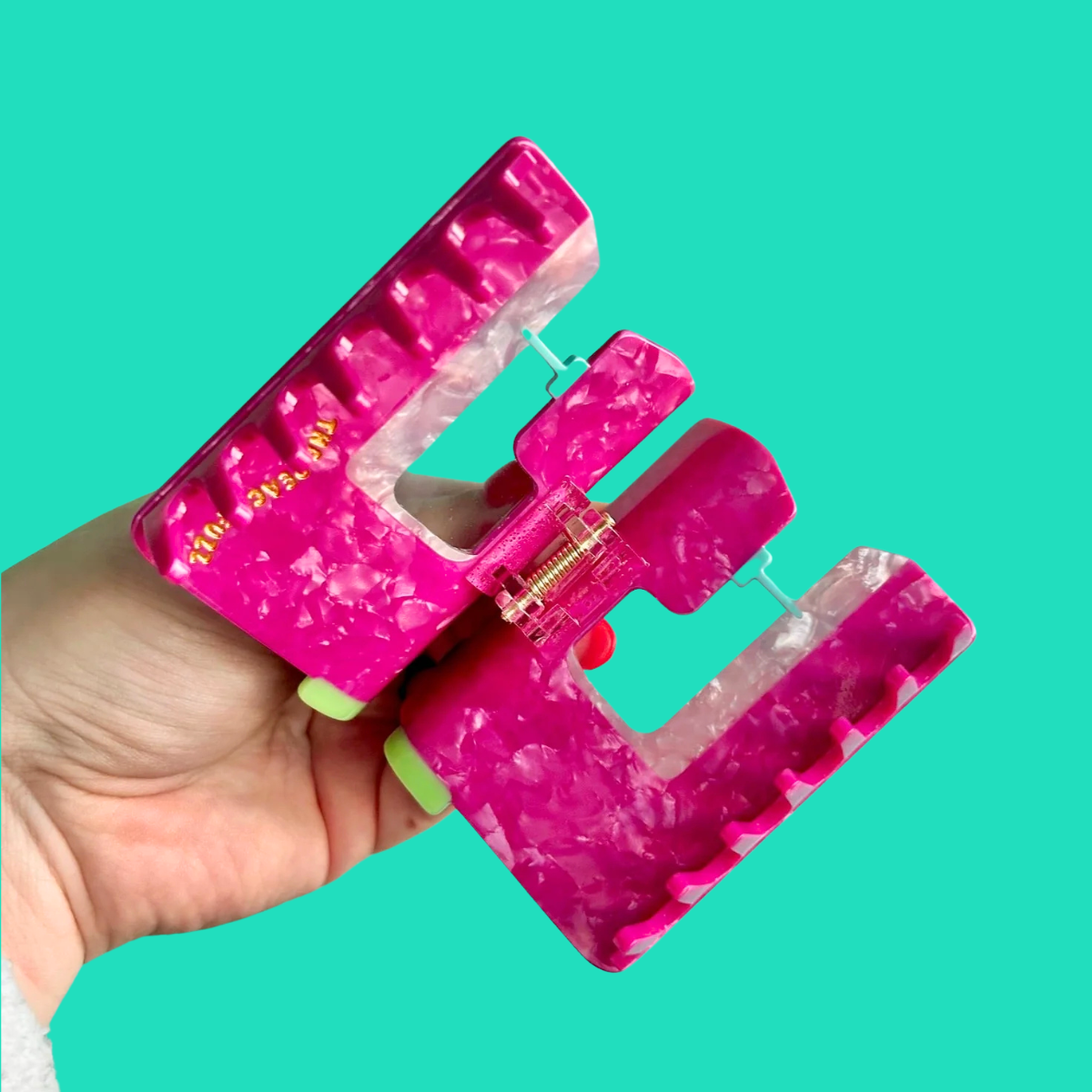 A hand holding a bright pink marbled claw hair clip against a solid turquoise background.