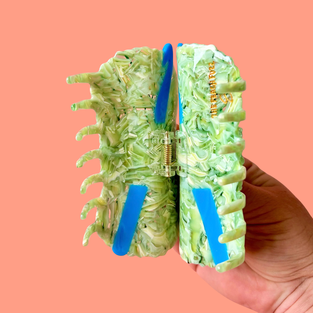A hand holds a large hair claw clip made from layered light green and white marbled material, with bright blue crochet hooks. The background is a solid peach-pink color.