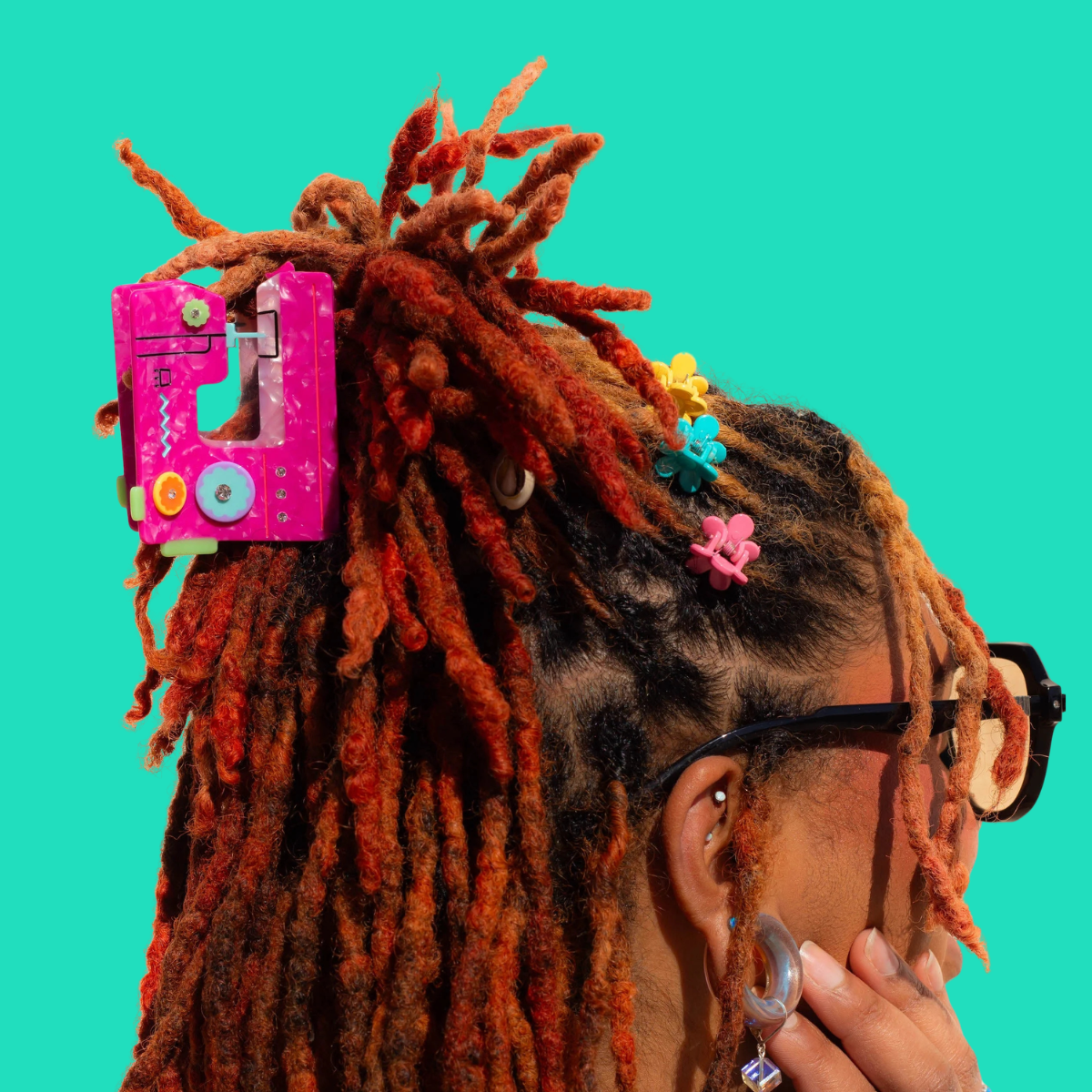 A close-up side profile of a person with long, textured orange-brown dreadlocks, wearing a bright pink sewing machine hair clip against a solid turquoise background.