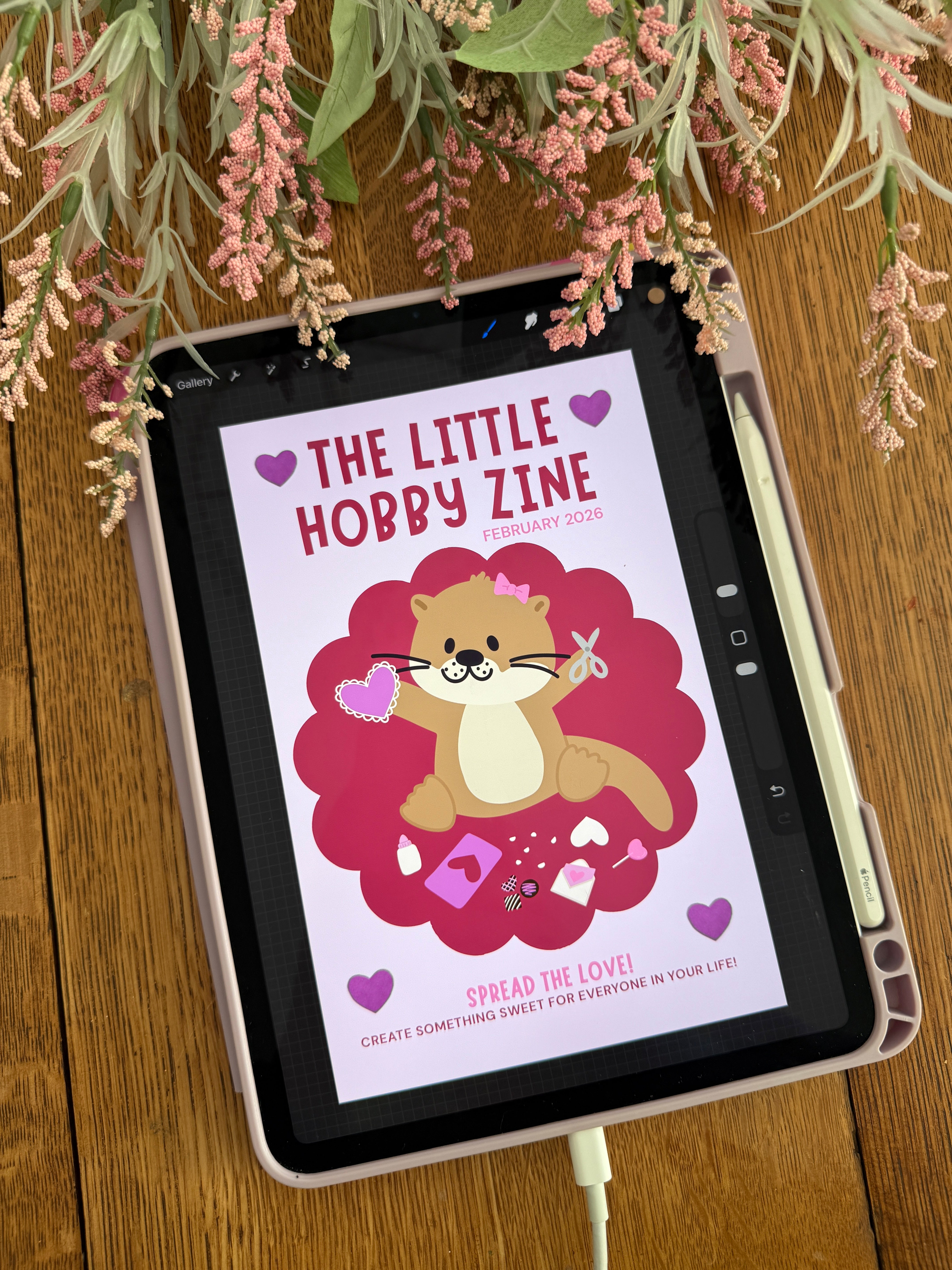 A iPad with a drawn pink and purple cover of The Little Hobby Zine. The cover has an otter crafting valentines. 