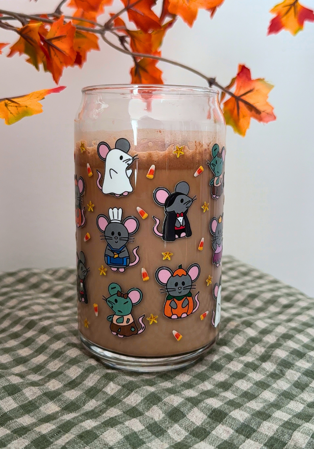 A clear glass can-shaped tumbler decorated with cute cartoon mice in Halloween costumes, filled with a brown beverage, set on a green and white checkered tablecloth with autumn leaves in the background.
