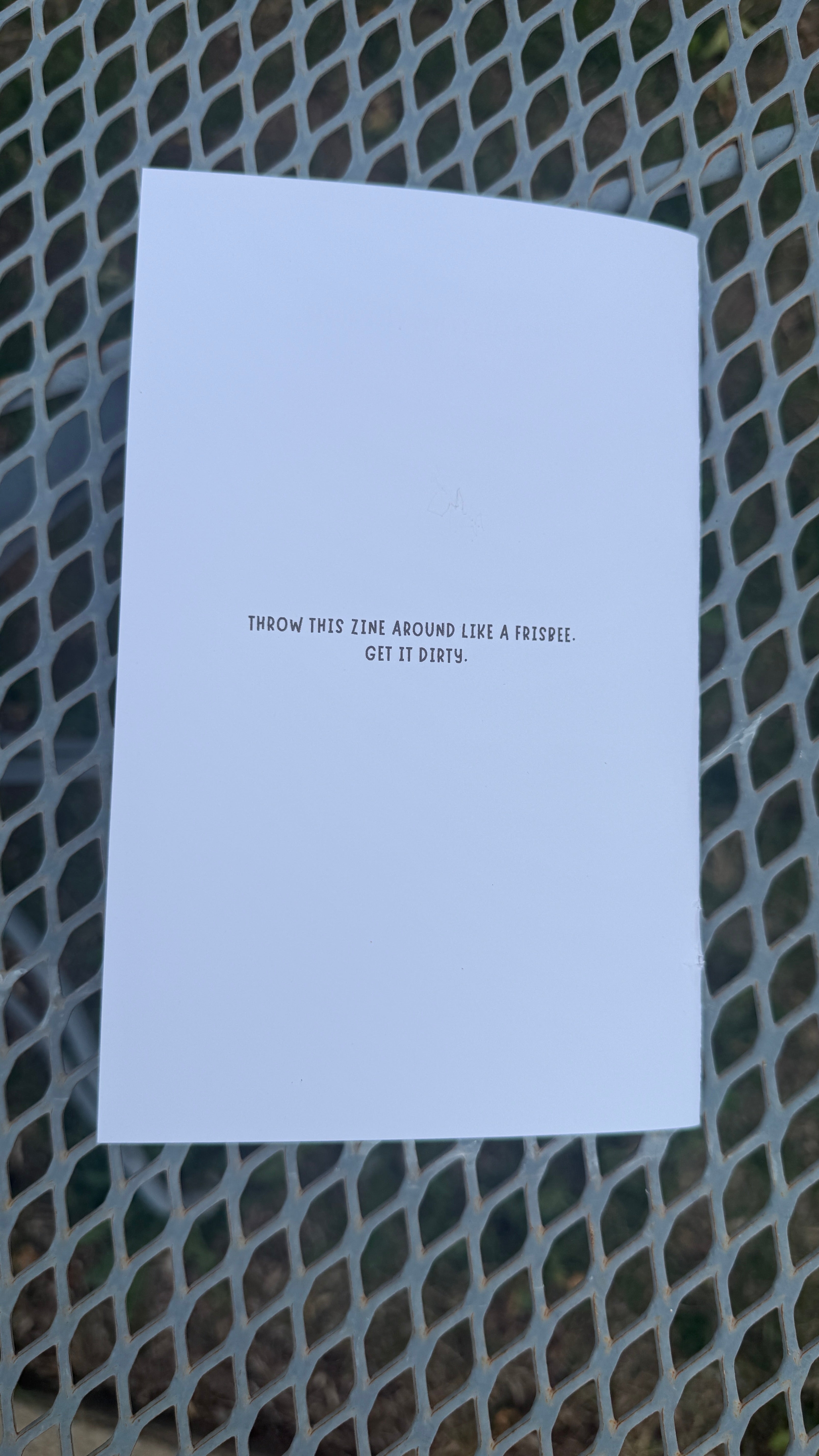 A white rectangular card or paper with small black text reading 'THROW THIS ZINE AROUND LIKE A FRISBEE. GET IT DIRTY.' placed on a gray metal mesh surface.