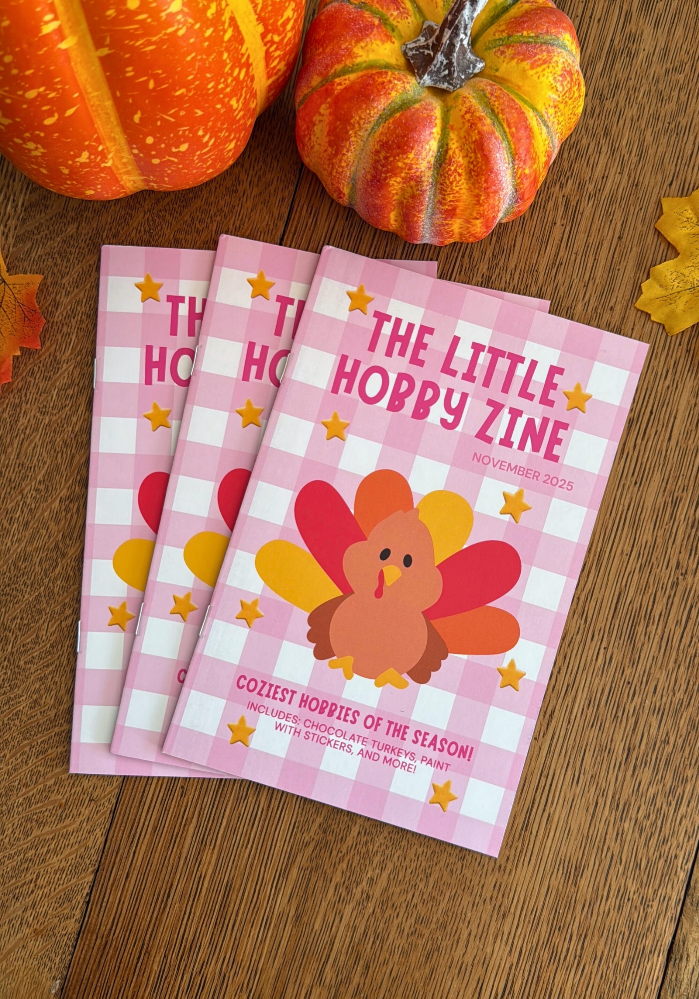 Three copies of 'The Little Hobby Zine' with a turkey illustration on a wooden surface with pumpkins.