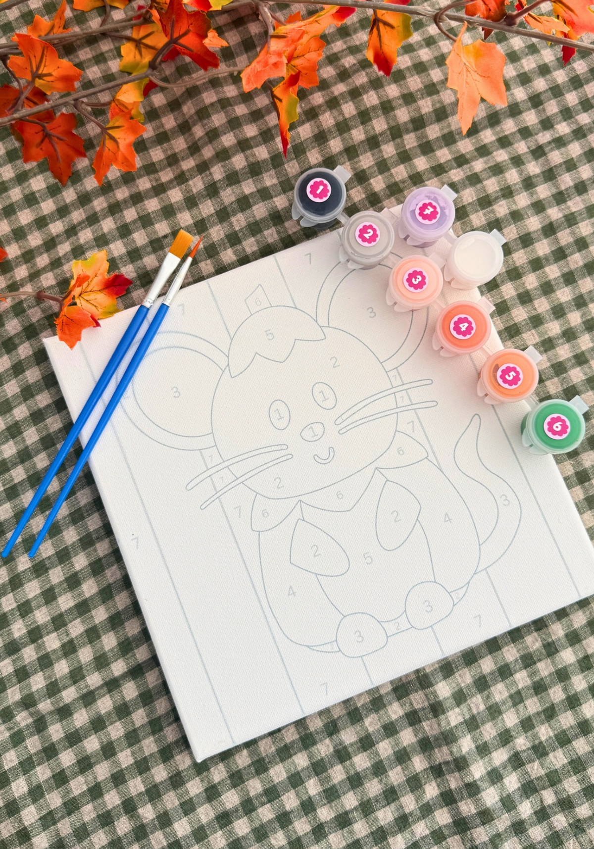 A canvas paint-by-numbers activity sheet featuring a cartoon cat design, styled on a green and white checkered blanket with autumn leaves. The sheet is surrounded by paint bottles in various colors and two blue paintbrushes.