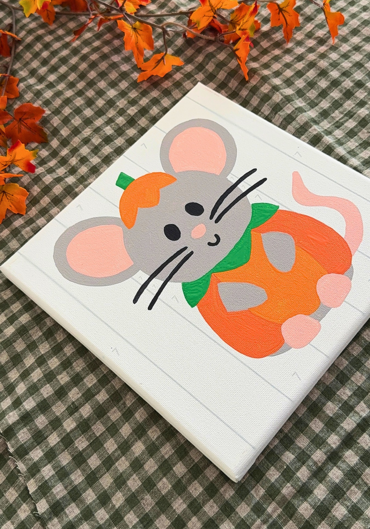A square canvas painting featuring a cute gray mouse wearing an orange pumpkin hat and a green scarf, holding an orange pumpkin. The canvas is placed on a green and white gingham checkered blanket with autumn leaves in the background.