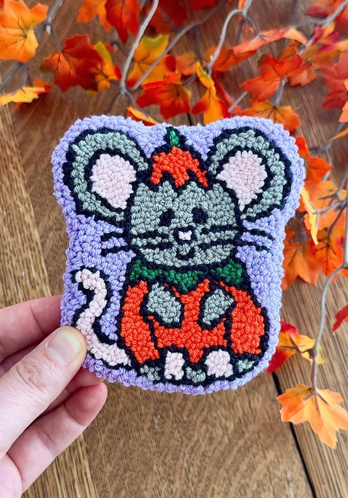 A hand tufted coaster of a mouse wearing a pumpkin costume on a light purple background.