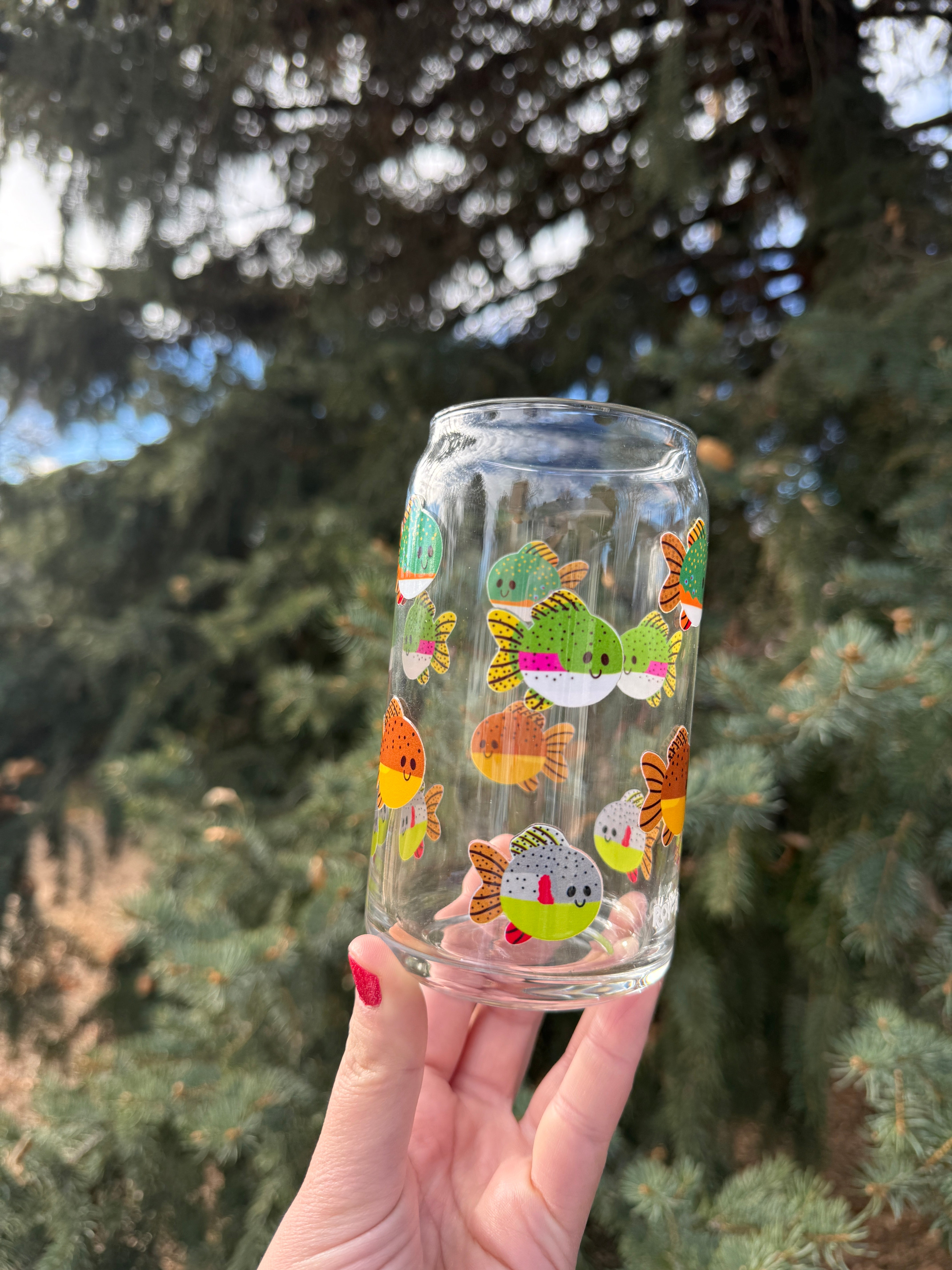 Clear glass with colorful trout design held by a hand against a blurred natural pine tree background