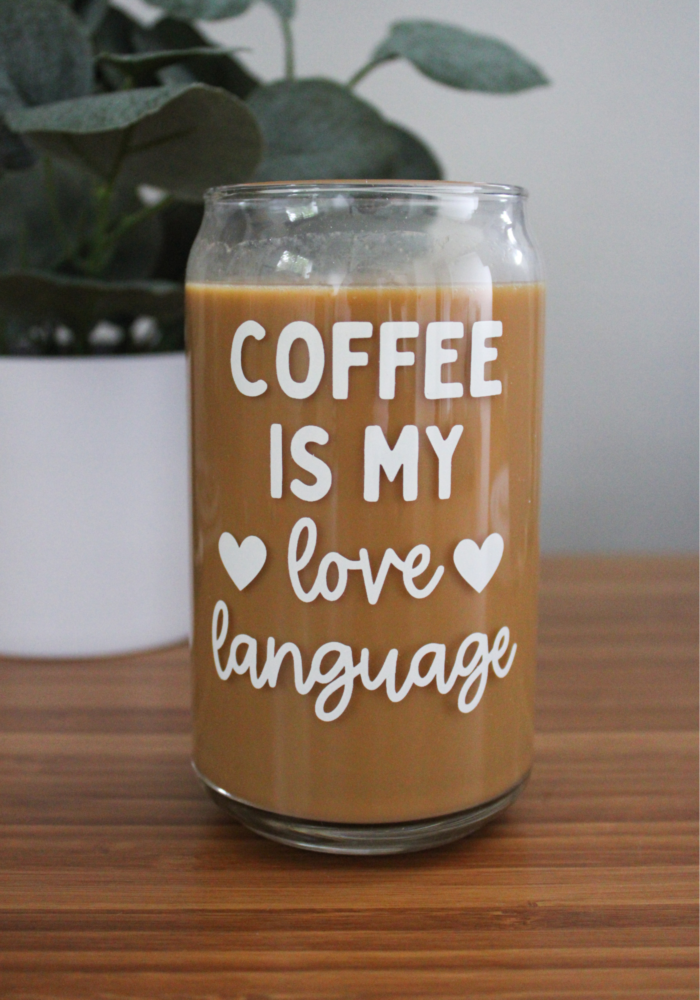 A clear glass coffee can with the text 'Coffee is my love language' printed on it, filled with coffee, placed on a wooden surface with a plant in the background.
