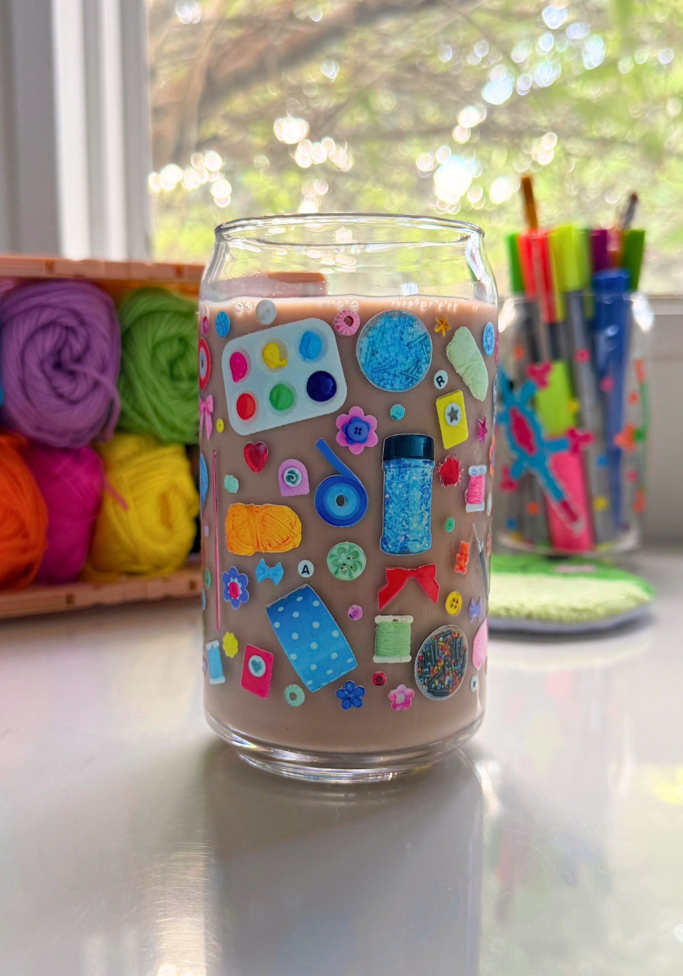 A clear glass can filled with chocolate milk, decorated with a colorful craft-themed decal, set on a reflective white surface with a window and craft supplies in the background.