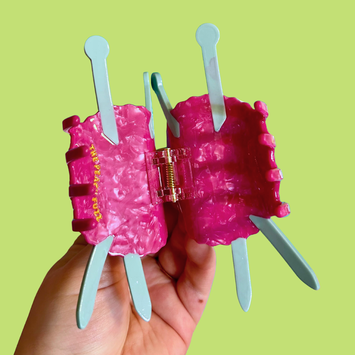 A hand holding a large, bright pink yarn hair claw clip with blue knitting needles, set against a solid light green background.