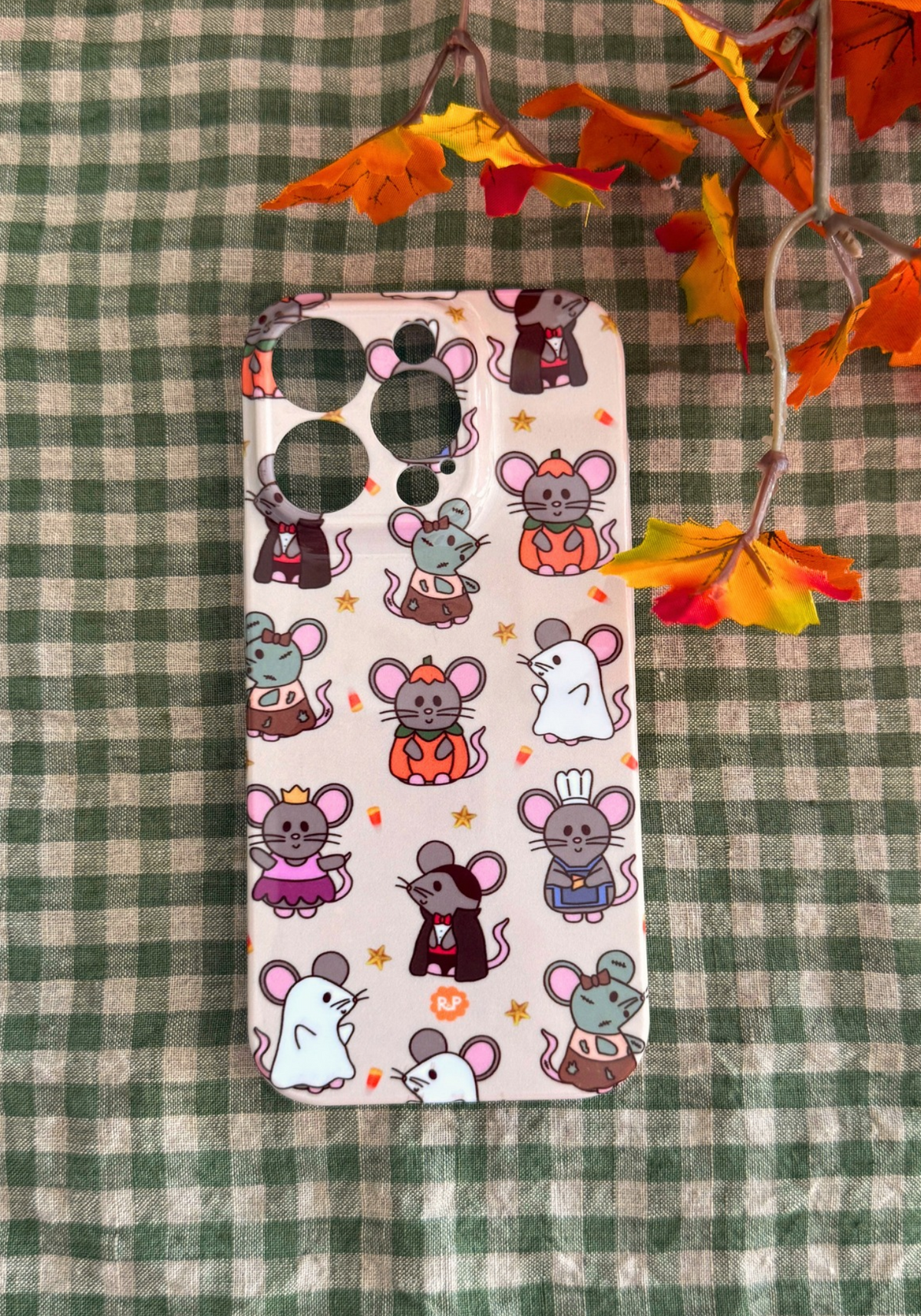 A phone case featuring a cute cartoon mouse pattern, laid flat on a green and white checkered blanket with decorative autumn leaves in the corner.