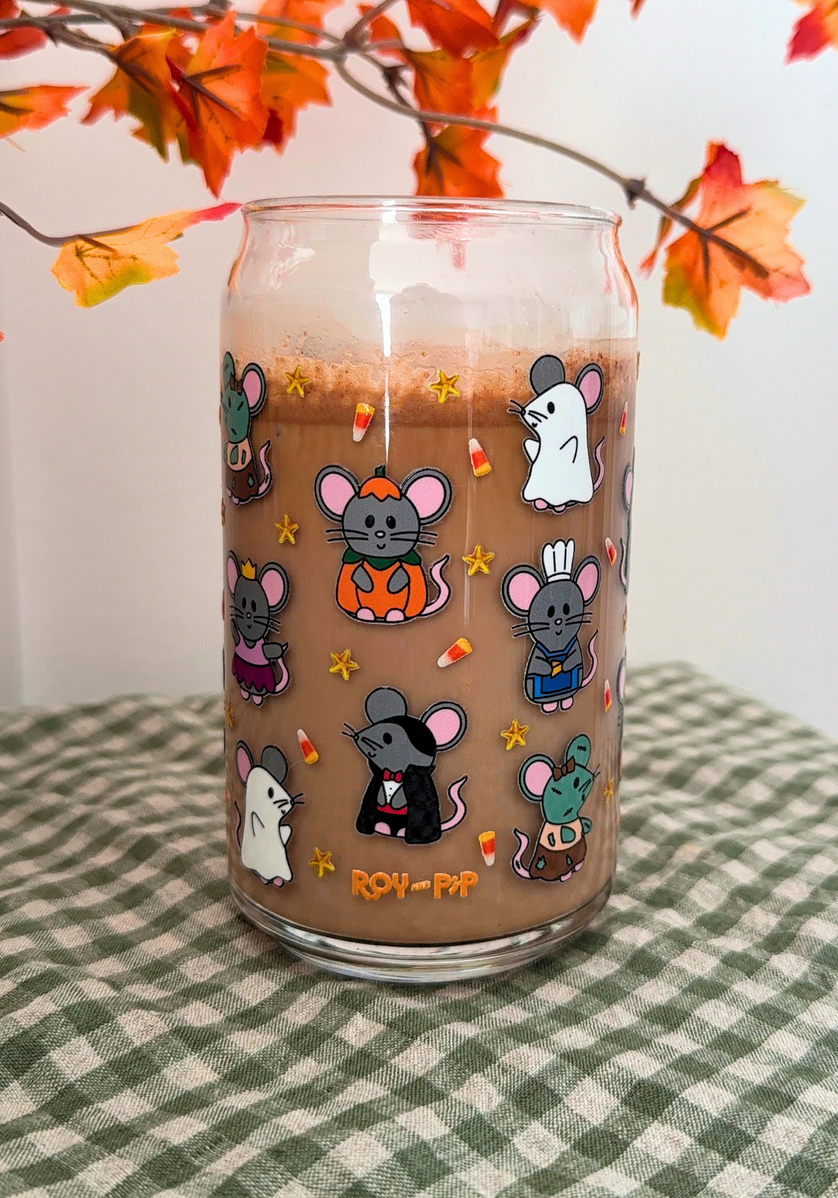 A clear glass can-shaped tumbler filled with a brown iced coffee or latte, decorated with cute cartoon mice in various outfits and small candy corn motifs. The glass is placed on a green and white checkered tablecloth with autumn maple leaves hanging in the background.