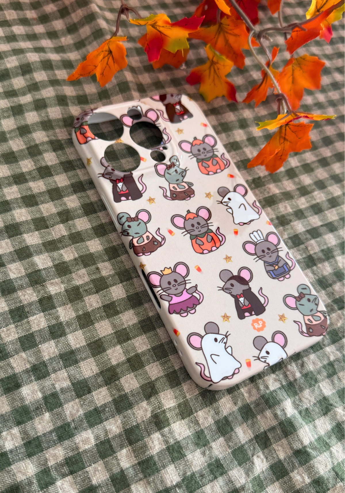 A phone case featuring a cute cartoon mouse pattern, displayed on a green and white checkered fabric background with decorative orange autumn leaves.